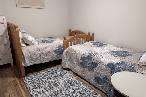 A bedroom features two wooden twin beds with blue floral bedding, a chevron-patterned rug, and a small round table. A framed 