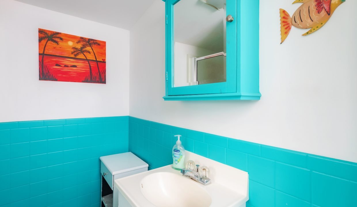 A bathroom with turquoise wall tiles, a white sink, a turquoise cabinet mirror, a soap dispenser, a fish wall decoration, and a sunset beach painting.