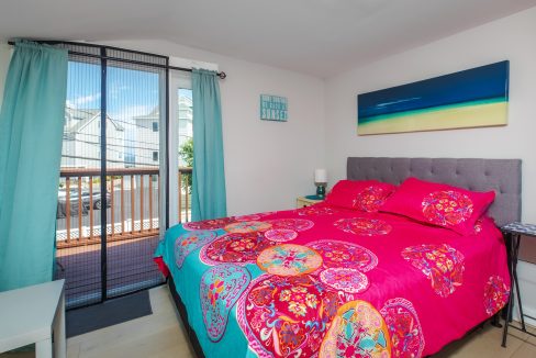 Bright bedroom with colorful bedding, a gray headboard, and a wall-mounted beach scene picture. Sliding glass doors with turquoise curtains lead to a small balcony outside.
