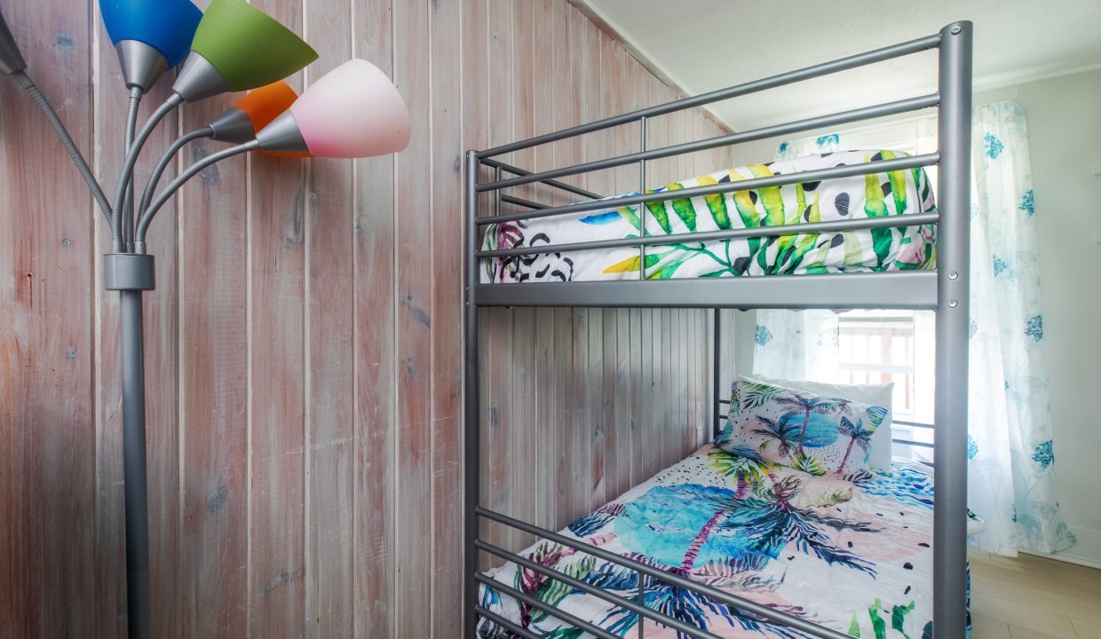 A small bedroom with a metal-framed bunk bed, colorful bedding, and a multi-colored floor lamp against a wooden wall. Natural light enters through a window with sheer curtains in the background.
