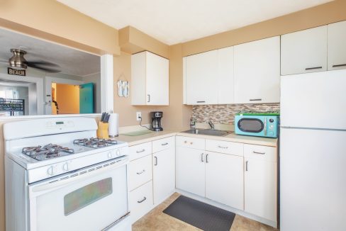 A modern kitchen with white cabinets, gas stove, coffee maker, turquoise microwave, and white refrigerator. A small backsplash is visible above the counter and sink area.