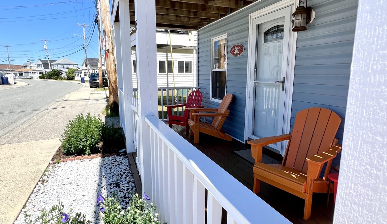 Porch with orange and red Adirondack chairs, white railing, and a flower bed with purple flowers in front of a light blue house under a clear blue sky.