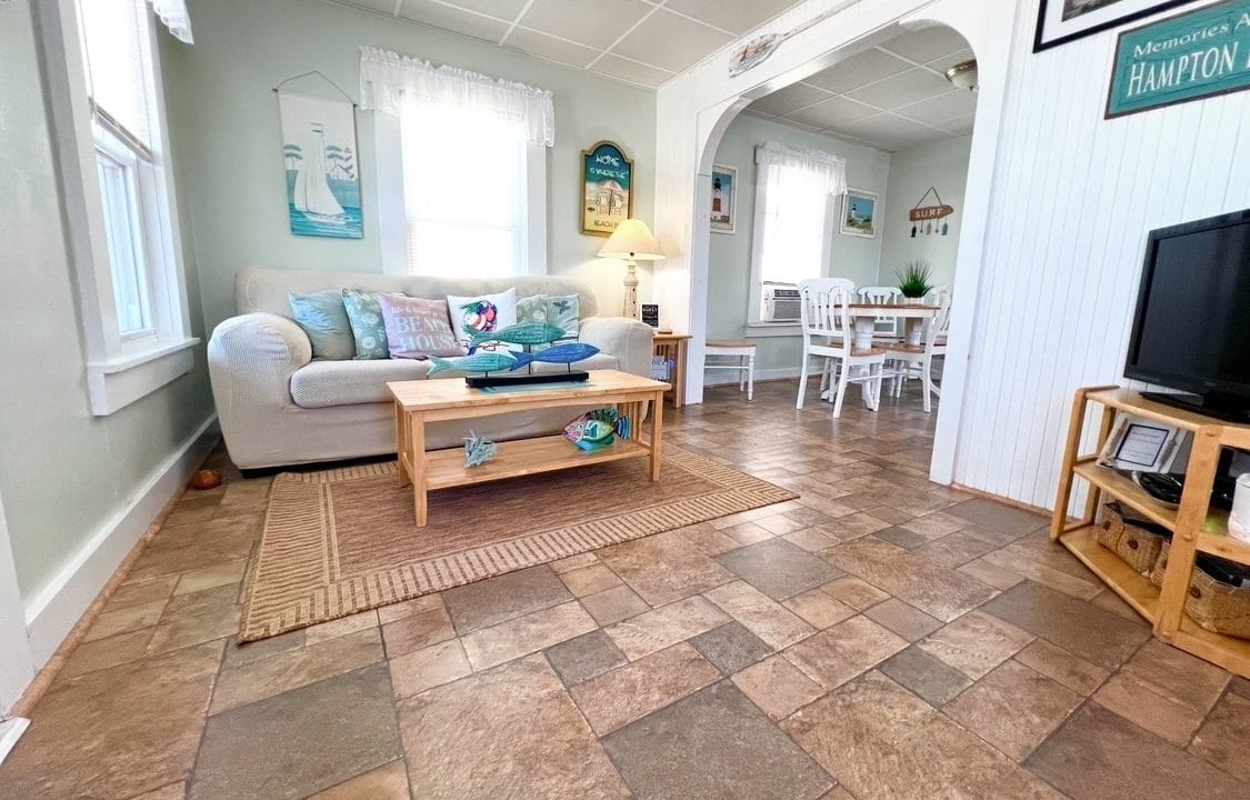 Cozy living room with a white couch, wooden coffee table, and a TV. Dining area with white chairs in the background. Decor features sailboat motifs.