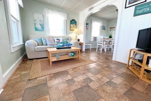 Cozy living room with a white couch, wooden coffee table, and a TV. Dining area with white chairs in the background. Decor features sailboat motifs.