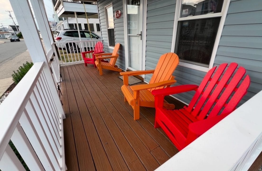 Three colorful Adirondack chairs in red and orange sit on a wooden porch outside a blue house.