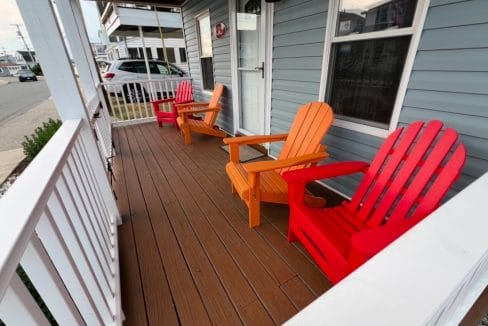 Three colorful Adirondack chairs in red and orange sit on a wooden porch outside a blue house.