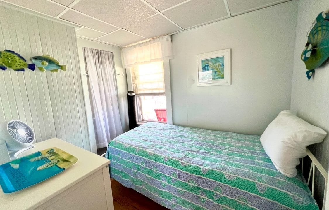 A small bedroom with a single bed covered in a blue patterned bedspread. Walls are white with fish decorations. There's a dresser with a fan and a tray, and a window in the back.