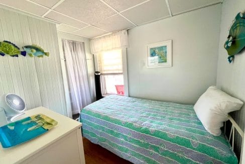 A small bedroom with a single bed covered in a blue patterned bedspread. Walls are white with fish decorations. There's a dresser with a fan and a tray, and a window in the back.