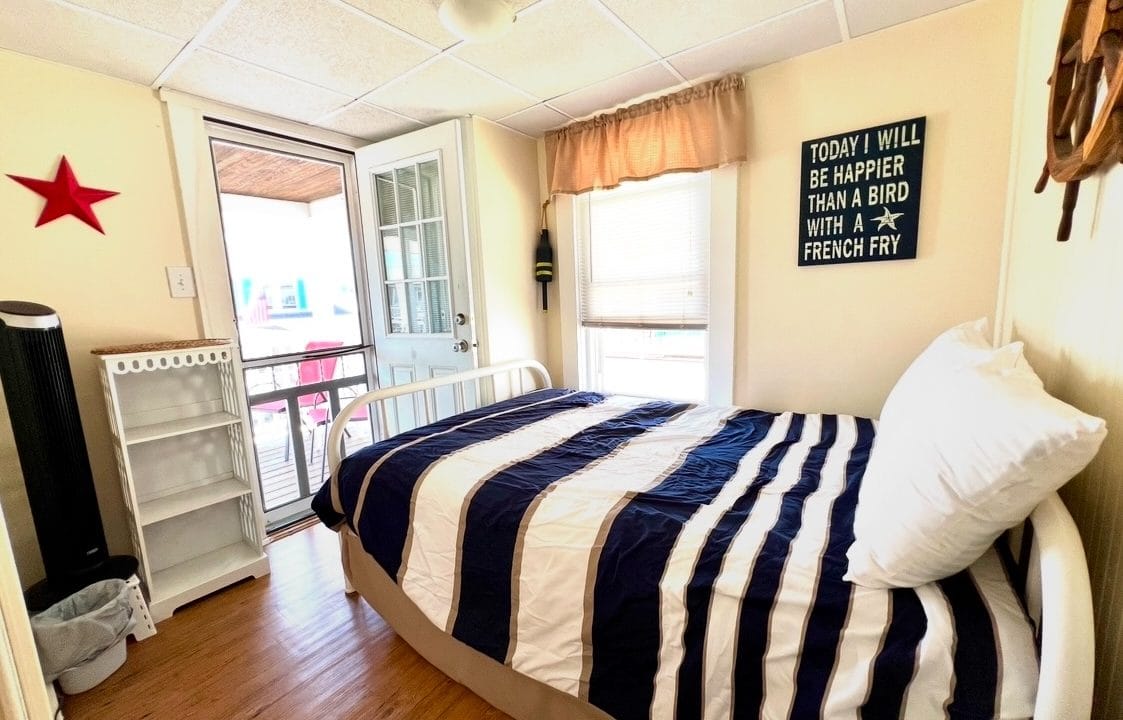 A small bedroom with a striped bedspread, a sign on the wall, and a window. The room has a door leading outside and light-colored walls, adorned with a red star decoration.