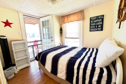 A small bedroom with a striped bedspread, a sign on the wall, and a window. The room has a door leading outside and light-colored walls, adorned with a red star decoration.