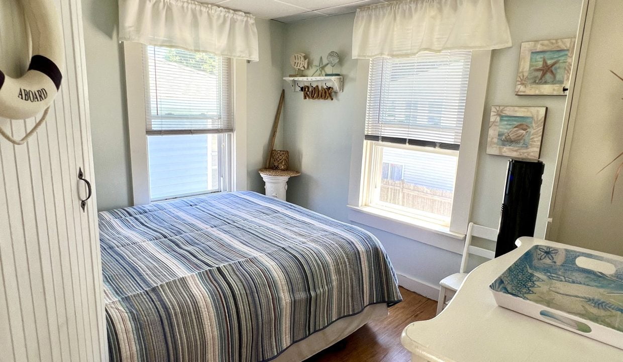 A small, cozy bedroom with a striped bedspread, white furniture, and coastal-themed decor, including wall art and a decorative lifebuoy. Two windows with white curtains allow natural light to enter.