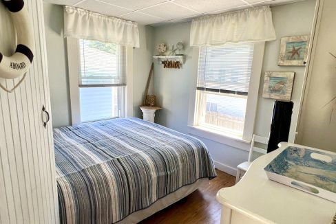 A small, cozy bedroom with a striped bedspread, white furniture, and coastal-themed decor, including wall art and a decorative lifebuoy. Two windows with white curtains allow natural light to enter.