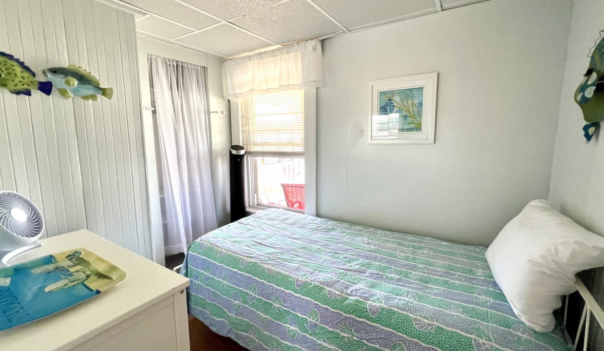 A small bedroom with a bed covered in a green and blue quilt, white dresser, and fish-themed wall decor. A window with sheer curtains brings in natural light.