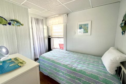 A small bedroom with a bed covered in a green and blue quilt, white dresser, and fish-themed wall decor. A window with sheer curtains brings in natural light.