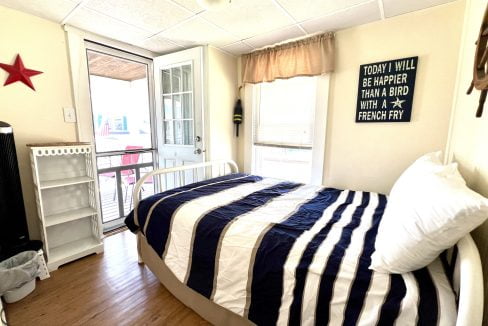 A small bedroom with a single bed covered in a black and white striped duvet, a mini fridge, a window, and a door leading to a balcony. A sign on the wall reads, 