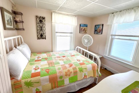 A small bedroom with a bed covered in a colorful patchwork quilt, two windows with white curtains, a fan on a white desk, and various wall decorations.
