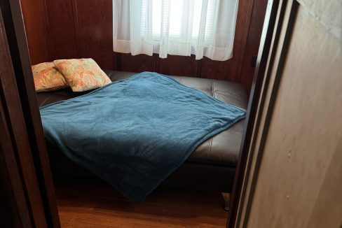 A small room with wooden walls and floor features a black couch bed with a blue blanket and two pillows near a window with sheer white curtains.