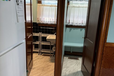 View of a kitchen area with a refrigerator on the left, leading to a laundry room with windows covered by curtains in the back. The floor is tiled, and wooden door frames separate the spaces.