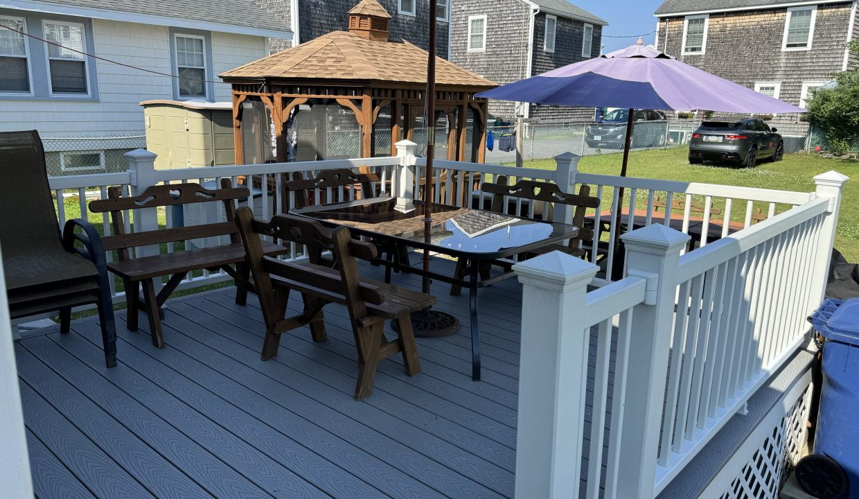 A backyard deck featuring wooden benches, a square table with folded chairs under a large patio umbrella, and a gazebo in the background. Nearby houses are visible.