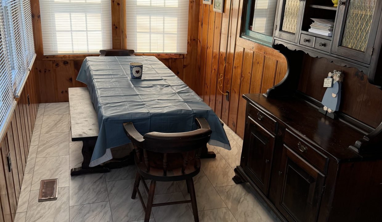 Wood-paneled dining room with a rectangular table covered with a blue tablecloth, surrounded by two benches and a chair. The room has three windows with patterned curtains and a dark wooden cabinet.