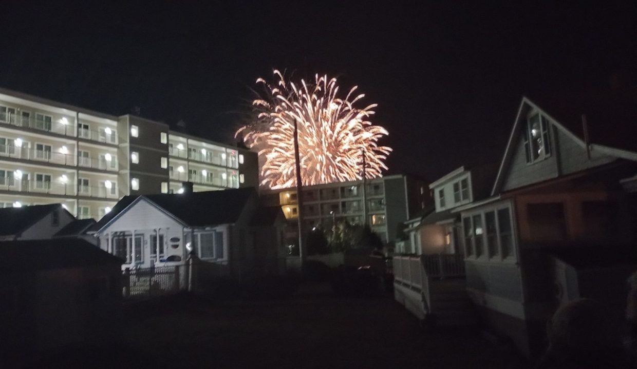 Fireworks light up the night sky behind a row of buildings and houses.