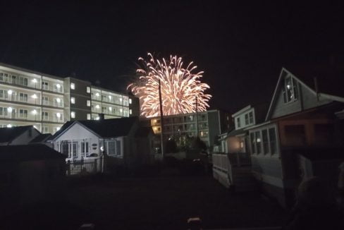 Fireworks light up the night sky behind a row of buildings and houses.