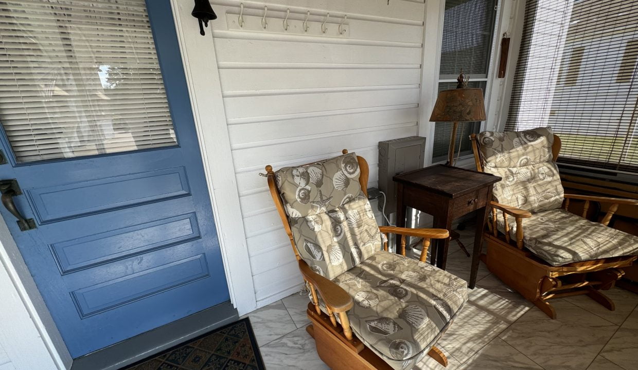 A small porch with two cushioned wooden chairs, a side table, and a blue door. A wall-mounted bell and a coat rack are also present. The area has wicker blinds and a tiled floor.