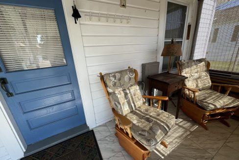 A small porch with two cushioned wooden chairs, a side table, and a blue door. A wall-mounted bell and a coat rack are also present. The area has wicker blinds and a tiled floor.