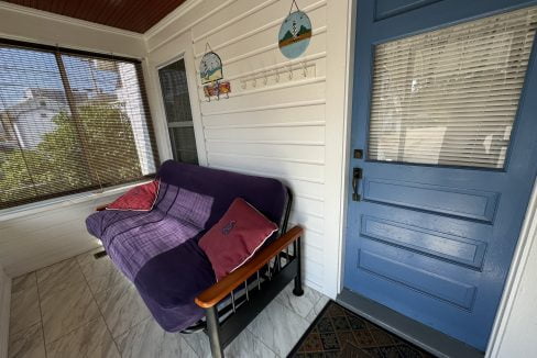 Enclosed porch with a blue door, a futon covered with a purple blanket, and decorative items on the wall. A window with closed blinds is on the left.