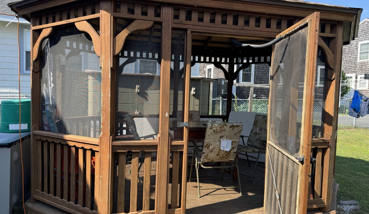 A wooden gazebo with a screen door and open sides, containing patio furniture, located in a backyard with neighboring houses visible in the background.