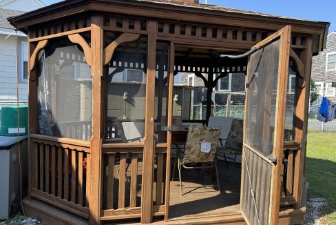 A wooden gazebo with a screen door and open sides, containing patio furniture, located in a backyard with neighboring houses visible in the background.