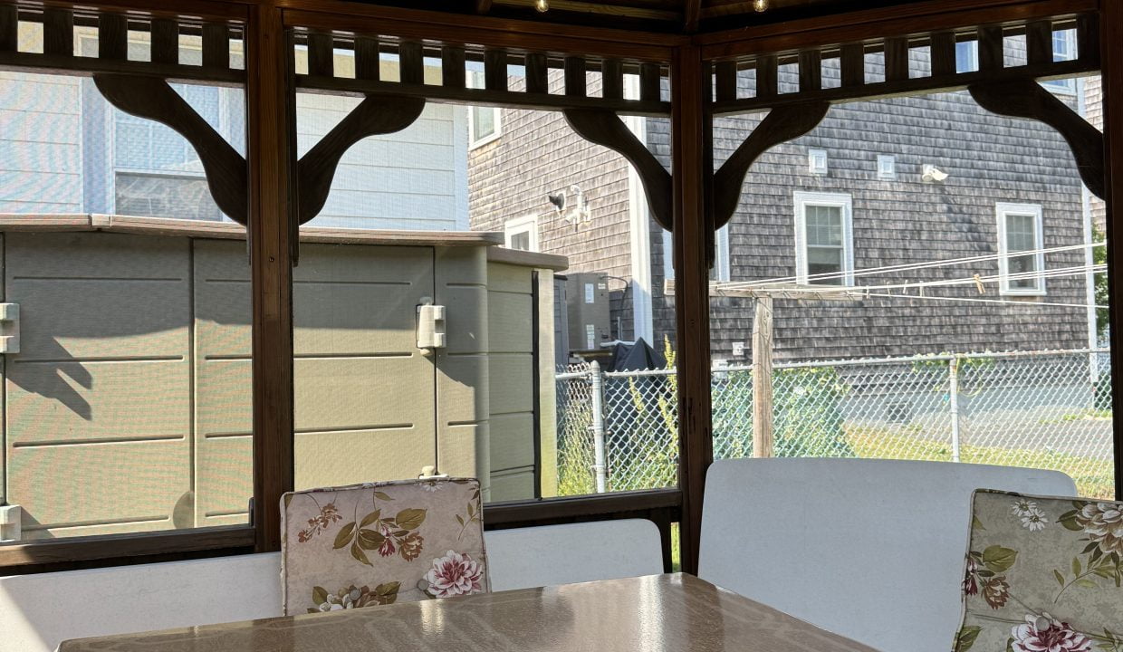 A gazebo with a wooden roof, containing a square table, floral-patterned chairs, and string lights. A television remote rests on the table. Background shows houses and a chain-link fence.