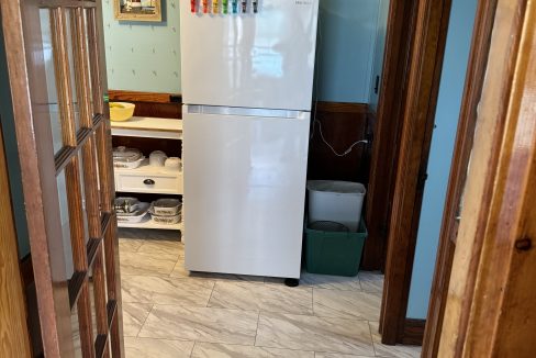 A white refrigerator in a room with light blue walls and a tiled floor. The fridge door has colorful magnets. Shelves to the left hold kitchen items, and a trash bin is to the right. Wall decor includes a lighthouse image.