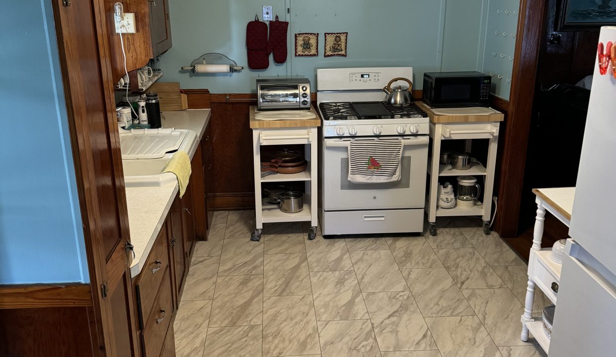 A small kitchen with wooden cabinets, a white stove, microwave, and fridge. The countertops hold various kitchen appliances, and the floor is tiled with a light gray pattern.
