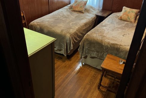 A small bedroom with two twin beds, a wooden nightstand between them, and a green dresser on the left. The room has wooden walls and a window with white curtains.