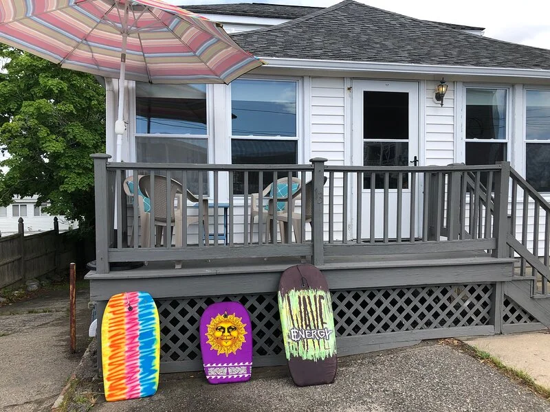 A wooden deck with three colorful bodyboards leaning against the railing in front of a house with a patio umbrella.