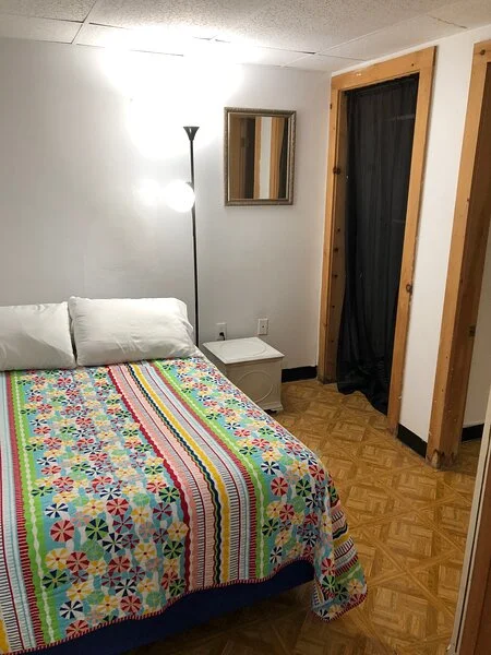 A modestly furnished bedroom with a patterned bedspread, floor lamp, and a mirror on the wall.