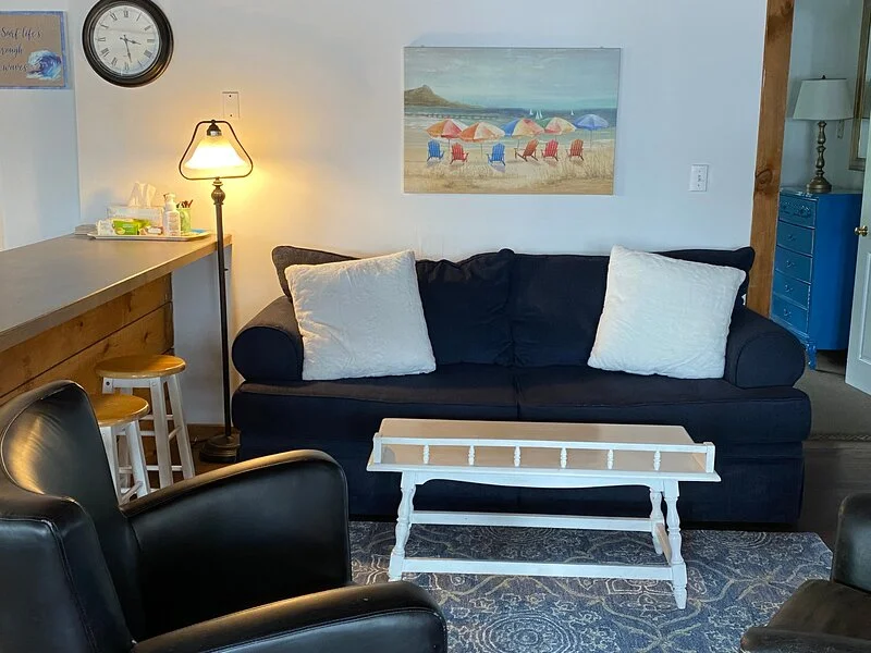 A cozy living room with a dark blue sofa, white pillows, a small white coffee table, and a beach painting on the wall.