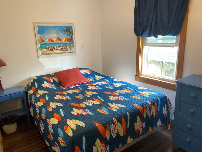 A tidy bedroom with a single bed featuring a colorful surfboard-themed blanket, a beach scene painting above the headboard, and coordinating blue furniture.