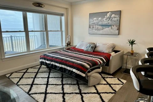 A neatly arranged bedroom with a striped comforter, a harbor-themed artwork, and a view of the water through large windows.