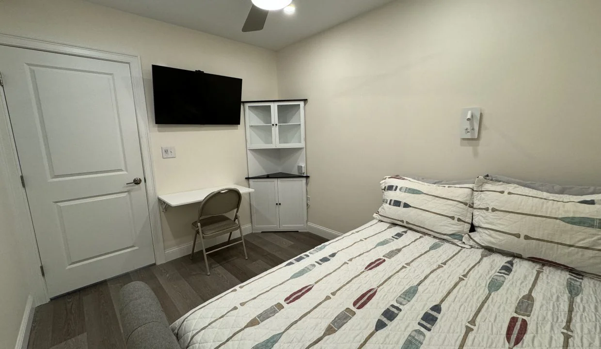 A small, neat bedroom with a bed covered in a patterned quilt, a wall-mounted tv, a desk with a chair, bookshelf, and a ceiling fan.