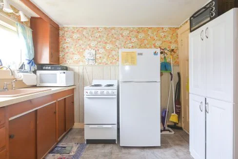 A small, vintage kitchen with floral wallpaper, wooden cabinets, and white appliances including a refrigerator, stove, and microwave.