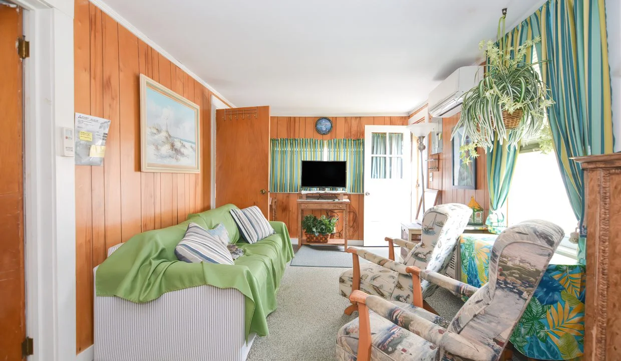 A cozy, well-lit living room with wooden paneling, floral curtains, and ample seating including a couch and chairs, a television set, and various houseplants.