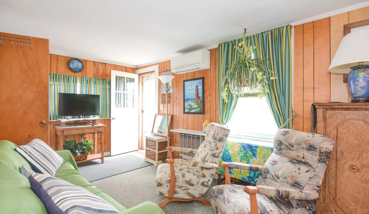 A brightly lit living room with green couch, patterned chairs, wood paneling, a television, and large window with curtains.