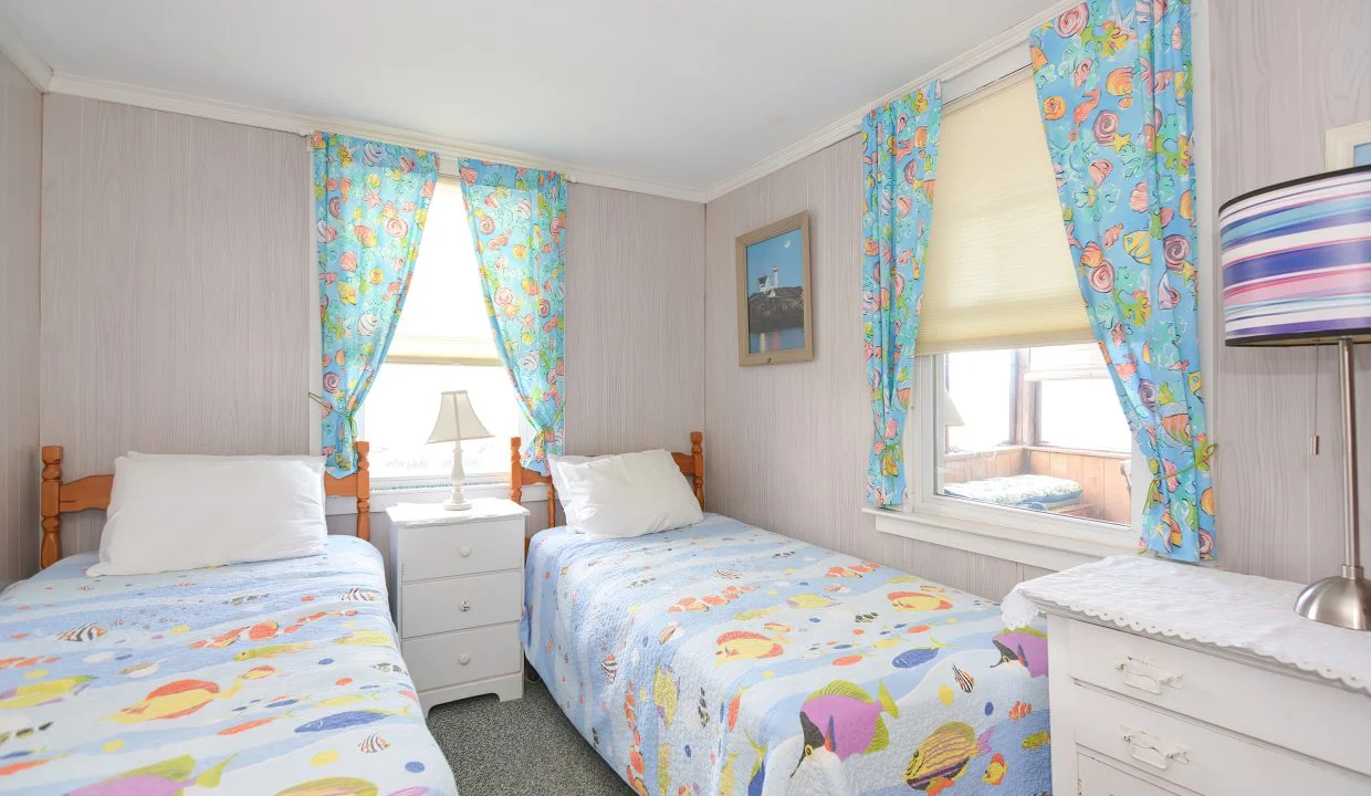 Bright, airy twin bedroom with colorful fish-themed bedding and curtains, white furniture, a small picture on the wall, and a visible window showing daylight.