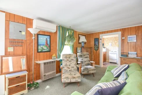 Interior of a vintage living room with wood paneling, patterned armchair, green sofa, curtain-draped windows, and doorway leading to another room.