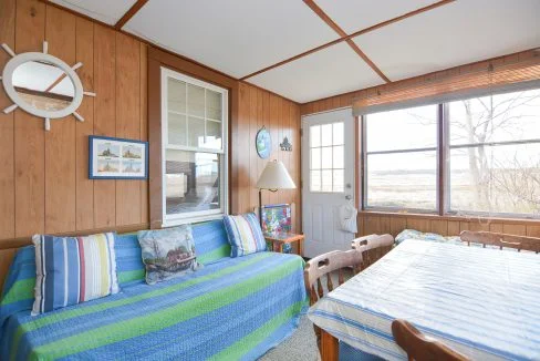 Cozy sunroom with wooden walls, large windows, patterned sofa, striped pillows, a round mirror, and a dining area with a wooden table.