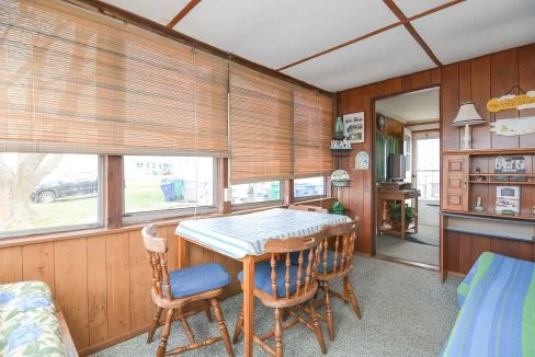 Interior of a cozy sunroom with wooden paneling, a table with chairs, blinds on large windows, and a display cabinet with various small items.