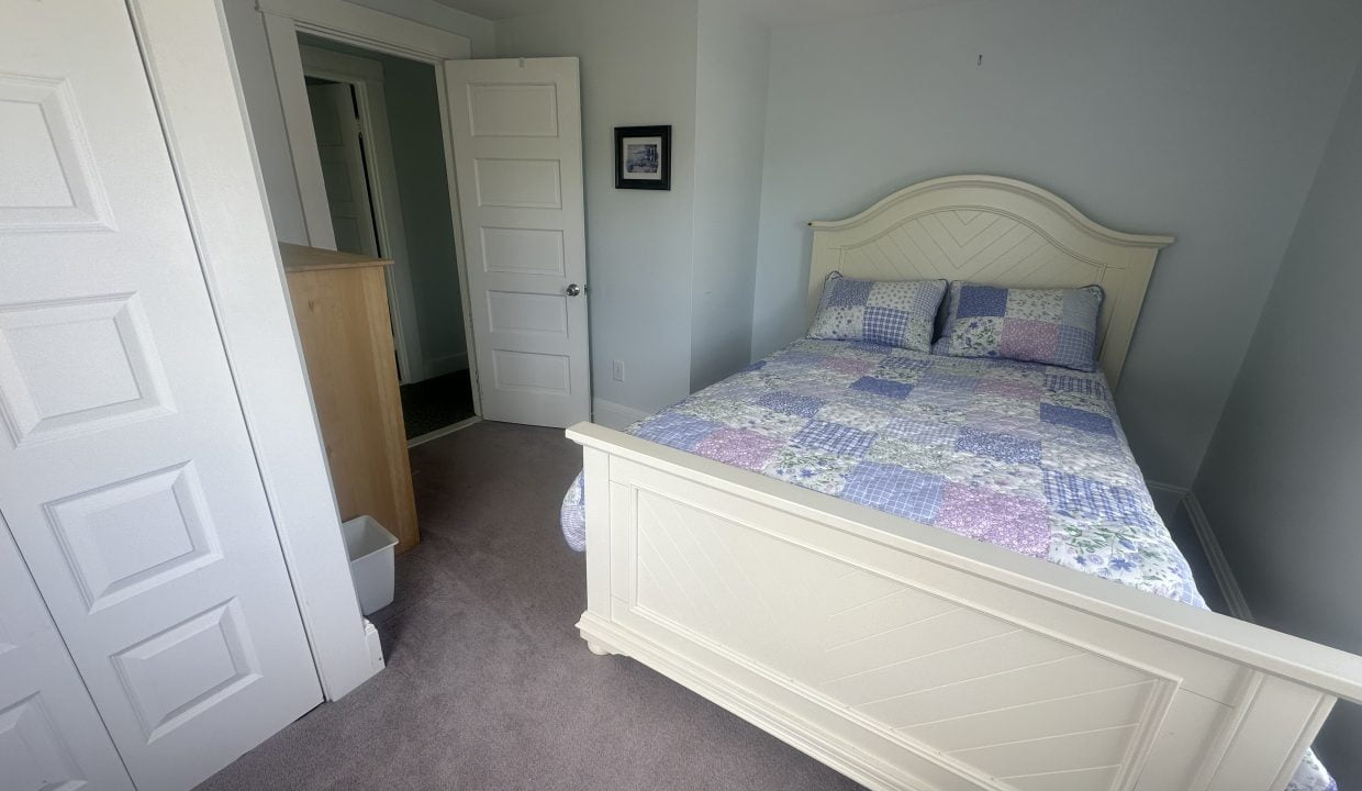 A small bedroom with a white bed frame and a patchwork quilt, a wooden dresser, a white door open to a hallway, and a small framed picture on the wall.