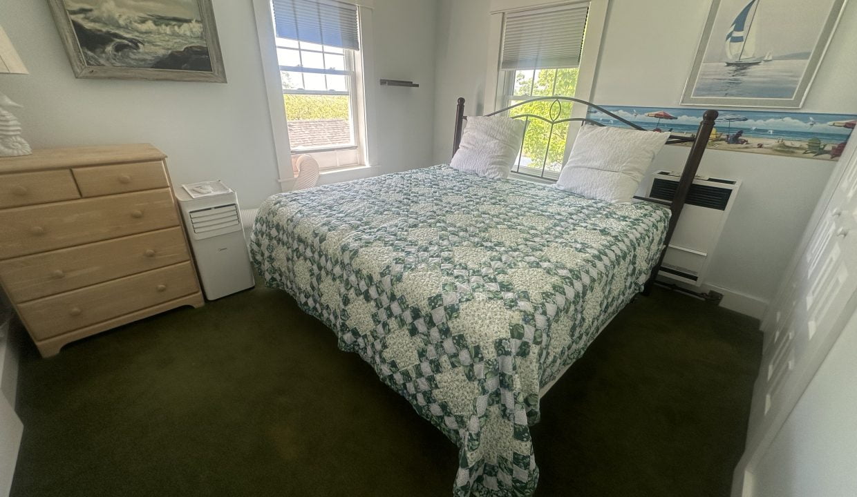 A small bedroom with a double bed covered in a green and white patterned quilt, a light wood dresser, and two windows with white blinds. A sailboat painting hangs on the wall above the bed.